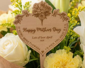May include: A bouquet of white roses, lilies, and yellow carnations wrapped in brown paper. A wooden heart-shaped sign with the text "Happy Mothers Day" and "I'd pick you first" is placed in the flowers.