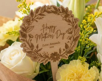 May include: A bouquet of white roses, lilies, and yellow carnations wrapped in brown paper. A wooden sign with the text "Happy Mother's Day" and "Love Amanda" is placed in the flowers.