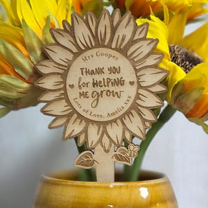 May include: A wooden sunflower plant stake with the text "THANK YOU for HELPING ME grow" and "Lots of Love, Amelia xxx". The stake is in a yellow vase with sunflowers.