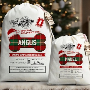 May include: Two cream-coloured Christmas gift sacks with drawstring closures. The larger sack is labelled "ANGUS" and the smaller sack is labelled "MABEL". Both feature a red and green plaid bone design, text, and festive stamps.