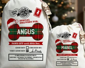 Puede incluir: Dos bolsas de regalo navideñas de color crema con cierres de cordón. La bolsa más grande está etiquetada como "ANGUS" y la más pequeña como "MABEL". Ambas presentan un diseño en forma de hueso a cuadros rojos y verdes, texto y sellos festivos.