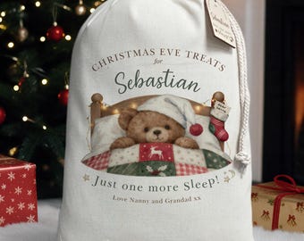 May include: A white fabric drawstring gift sack featuring a printed illustration of a teddy bear in a bed with a quilt. The text reads: CHRISTMAS EVE TREATS for Sebastian, Just one more Sleep!, and Love Nanny and Grandad xx. A small tag is attached.
