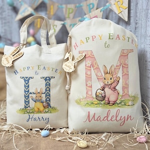 May include: Two off-white Easter-themed tote bags and drawstring bags. The bags feature the text "Happy Easter to" and a letter, along with a cartoon bunny. One bag says "Harry" and the other says "Madelyn". Each bag has a wooden bunny tag.