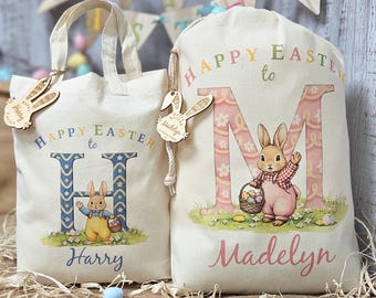 May include: Two off-white Easter-themed tote bags and drawstring bags. The bags feature the text "Happy Easter to" and a letter, along with a cartoon bunny. One bag says "Harry" and the other says "Madelyn". Each bag has a wooden bunny tag.