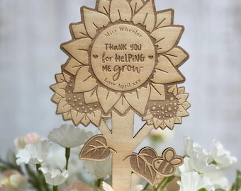 May include: A wooden sunflower-shaped decoration with the text "THANK YOU for HELPING ME grow" and the name "Miss Wheeler". The decoration has a tree trunk and a bee and leaf design. The decoration is placed in a bouquet of flowers.