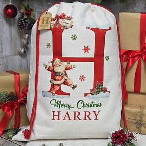 May include: A white Christmas gift bag with a large red letter "H" and a Santa Claus illustration. The bag has a red drawstring and the words "Merry Christmas HARRY". Presents wrapped in brown paper with red ribbons are in the background.