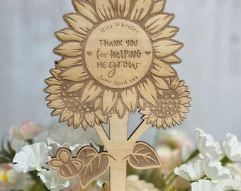 May include: Wooden sunflower-shaped teacher appreciation gift. The design includes a sunflower, tree trunk, leaves, and a bee. The text reads "Miss Wheeler, THANK YOU for HELPING ME grow, Love April xxx."