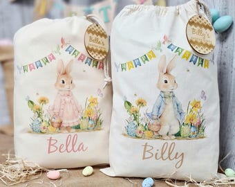 May include: Two personalized Easter gift bags with bunny illustrations. The bags are made of natural fabric and feature a drawstring closure. One bag is decorated with a pink bunny and the name "Bella" and the other with a blue bunny and the name "Billy".