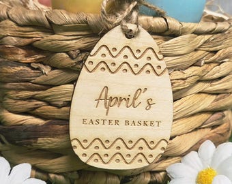 May include: A woven Easter basket with colorful eggs and a wooden egg-shaped tag. The tag is engraved with the text "April's EASTER BASKET" and decorative wave and dot patterns. The basket is decorated with daisies.