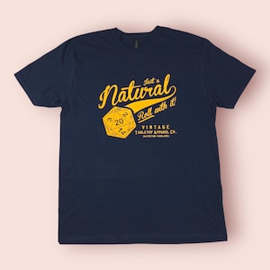 May include: Navy blue t-shirt with a yellow graphic featuring a twenty-sided die and the text "Just a Natural Roll with it!". The shirt also includes "Vintage Tabletop Apparel Co." and the location "(Selfeton - England)".