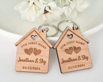 May include: Two house-shaped wooden keychains featuring a metal ring. Each keychain is engraved with the text "OUR FIRST HOME", two hearts, the names "Jonathan & Sky", and the date "21/12/2024". The design is consistent on both items.
