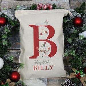 Puede incluir: Una bolsa de regalo navide&ntilde;a de color crema con una gran letra "B" roja con un mu&ntilde;eco de nieve y copos de nieve. La bolsa dice "Merry Christmas BILLY" en una fuente festiva. La bolsa est&aacute; rodeada por una corona navide&ntilde;a con adornos rojos.