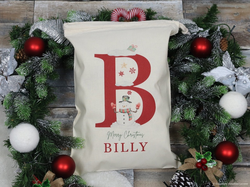 Puede incluir: Una bolsa de regalo navide&ntilde;a de color crema con una gran letra "B" roja con un mu&ntilde;eco de nieve y copos de nieve. La bolsa dice "Merry Christmas BILLY" en una fuente festiva. La bolsa est&aacute; rodeada por una corona navide&ntilde;a con adornos rojos.