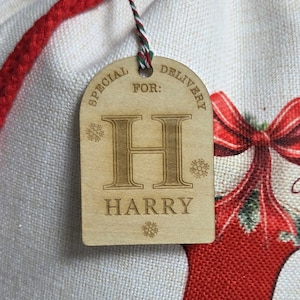 May include: A wooden gift tag with the text "SPECIAL DELIVERY FOR: HARRY". The tag is light brown with engraved text and snowflake designs. It hangs from a red and green string, with a red drawstring visible in the background.