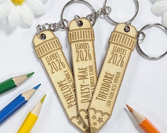 May include: Three wooden pencil-shaped keychains with silver keyrings. Each keychain is engraved with "LEAVERS 2026" and a name: Penelope, Daisy-Mae, and Theodore. Colored pencils in green, blue, yellow, and orange are in the background.