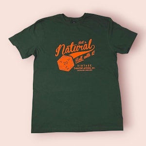 May include: Dark green t-shirt with an orange graphic that reads "Just a Natural Roll with it!" and a die. The shirt also has the text "Vintage Tabletop Apparel Co." below the die.