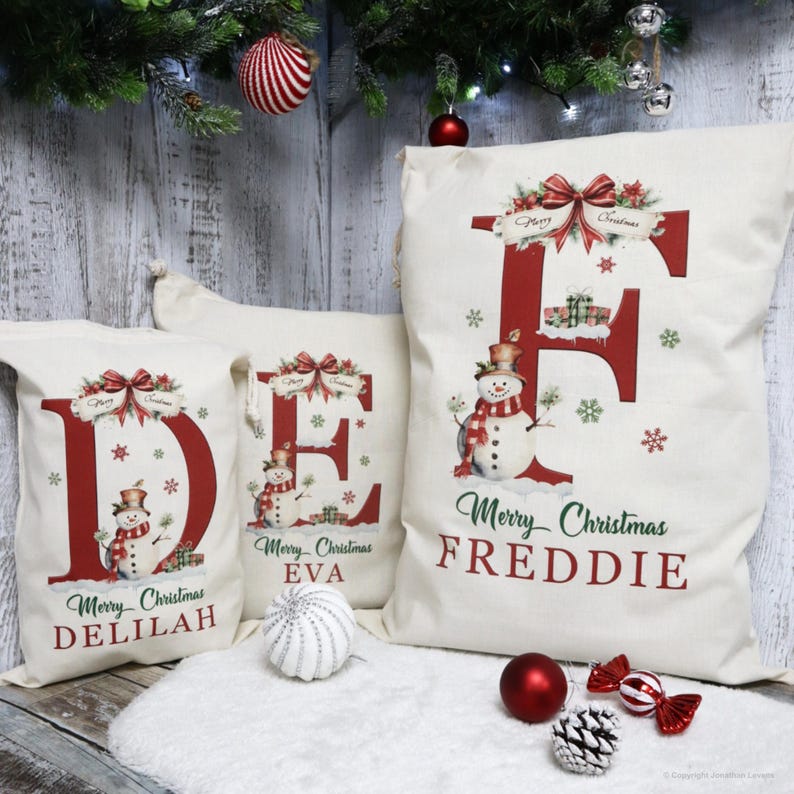 May include: Three off-white Christmas gift bags with red lettering and snowman illustrations. The bags feature the letters "D", "E", and "F", with the names "Delilah", "Eva", and "Freddie" printed below. Festive holiday decor.