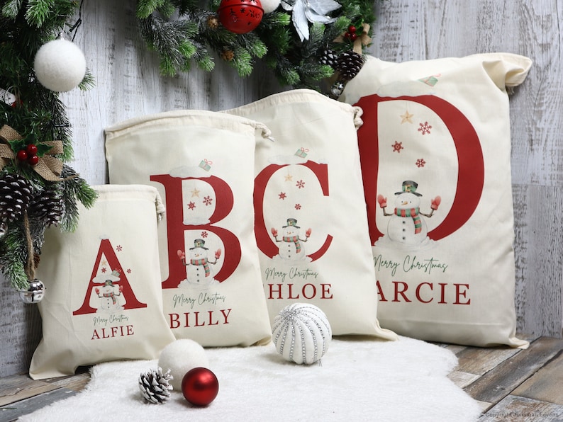 Puede incluir: Cuatro bolsas de regalo navide&ntilde;as color crema con letras rojas A, B, C y D. Cada bolsa presenta un mu&ntilde;eco de nieve, copos de nieve y la frase "Merry Christmas". Los nombres est&aacute;n impresos debajo. Decoraci&oacute;n navide&ntilde;a festiva.