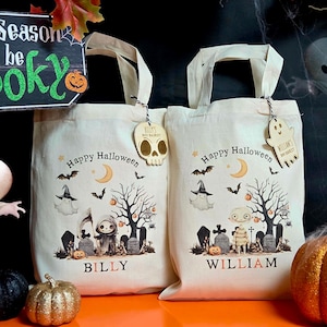 May include: Two cream-colored Halloween tote bags with spooky illustrations, including ghosts, bats, and tombstones. Each bag has a wooden tag and the words "Happy Halloween." One bag says "Billy" and the other says "William."