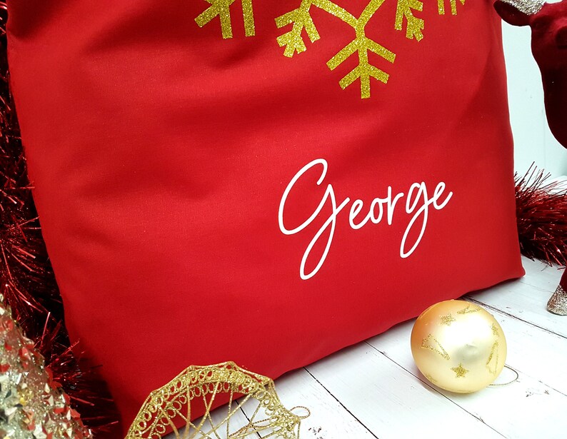 May include: Red fabric gift bag with a gold snowflake design and the name "George" printed in white.