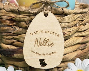 May include: A wooden Easter egg tag with the words "Happy Easter Nellie" engraved on it, along with a bunny silhouette. The tag is tied to a woven basket containing decorated Easter eggs.