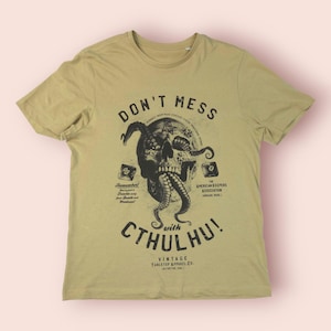 May include: A khaki-colored t-shirt featuring a black graphic of a skull with tentacles, and the text "DON'T MESS WITH CTHULHU!". The shirt also includes additional text and design elements, with a vintage aesthetic.