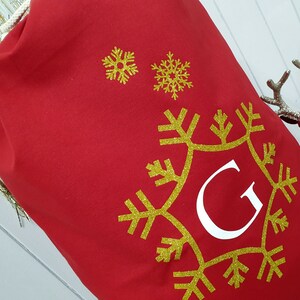 May include: Red Christmas gift bag with a gold snowflake design and the letter "G" in white.