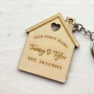 May include: A wooden house-shaped keyring with a heart-shaped cut-out. The keyring is engraved with the words "OUR FIRST HOME" above "Tansy & Tyler" and "EST. 10/11/2023". A silver keyring is attached.