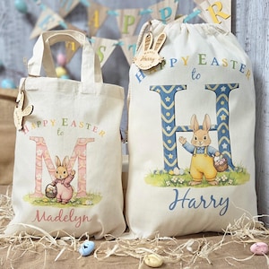 May include: Two off-white Easter tote bags. One bag has the text "Happy Easter to M" with a bunny and the name "Madelyn". The other bag has the text "Happy Easter to Harry" with a bunny and the letter "H". Both bags have wooden bunny-shaped tags.