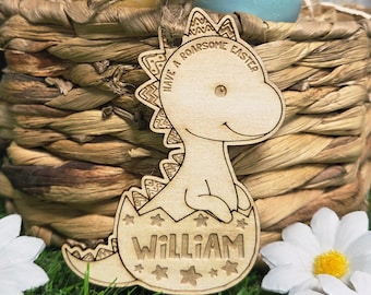 May include: A wooden dinosaur-shaped Easter ornament with the text "Have a Roarsome Easter" and the name "William". The ornament is light brown and has a decorative edge. It is placed in front of a woven basket with Easter eggs.