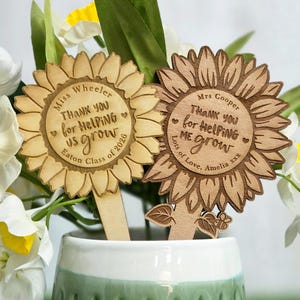 May include: Two wooden sunflower-shaped plant markers with engraved text. The left marker reads Miss Wheeler THANK YOU for HELPING us grow Eaton Class of 2026. The right marker reads Mrs Cooper THANK YOU for HELPING ME grow Lots of Love, Amelia xxx.