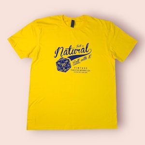 May include: A vibrant yellow t-shirt featuring a navy blue graphic. The design includes the phrase "Just a Natural Roll with it!" and a die showing the number 20. From Vintage Tabletop Apparel Co.