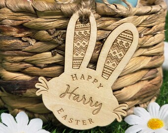 May include: A wooden Easter bunny tag with the name "Harry" engraved, along with the words "Happy Easter." The bunny ears have a decorative pattern. The tag is attached to a woven basket containing colorful Easter eggs, with white daisies in the foreground.