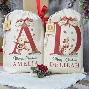 May include: Two cream-coloured Christmas gift bags with red lettering and illustrations. One bag features a red "A" with Santa Claus, the other a red "D" with a snowman. Both bags have "Merry Christmas" and a name printed below.