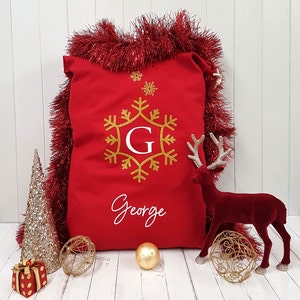 May include: Red personalized Christmas stocking with a gold snowflake and the name George. The stocking is surrounded by gold tinsel and a red reindeer figurine.