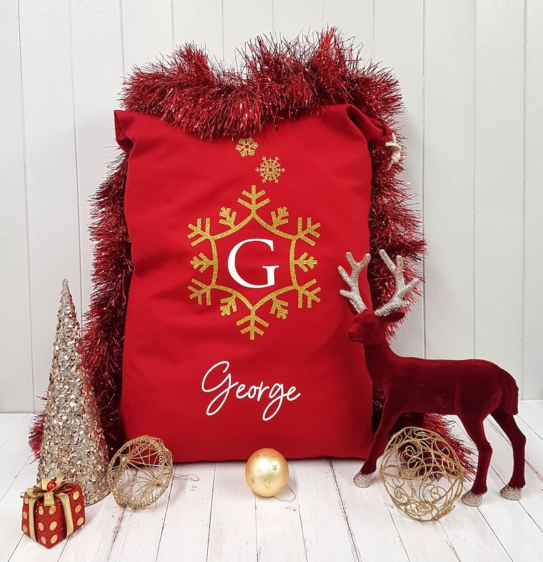 May include: Red personalized Christmas stocking with a gold snowflake and the name George. The stocking is surrounded by gold tinsel and a red reindeer figurine.