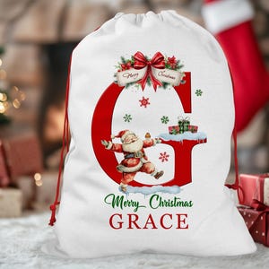 May include: White Christmas gift bag with a red drawstring. The bag features a large red letter "G" with a Santa Claus illustration, snowflakes, and the text "Merry Christmas GRACE". A red bow and greenery adorn the top.