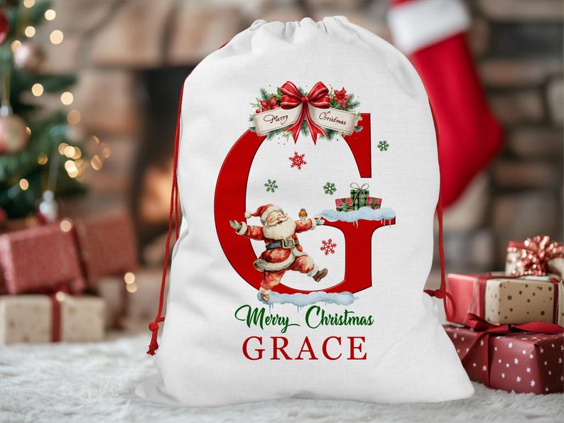 May include: White Christmas gift bag with a red drawstring. The bag features a large red letter "G" with a Santa Claus illustration, snowflakes, and the text "Merry Christmas GRACE". A red bow and greenery adorn the top.