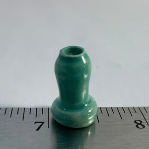 Op de afbeelding: Een kleine, teal-kleurige keramische vaas met een ronde basis en een licht uitlopende opening. De vaas is ongeveer 2,5 cm hoog en staat op een liniaal.