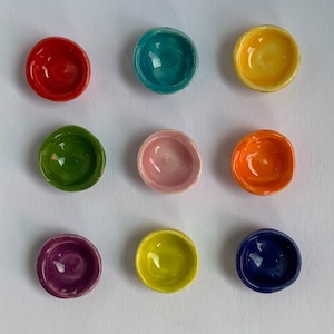 Pode incluir: Nove pequenas tigelas de cerâmica em várias cores, incluindo vermelho, verde, azul, amarelo, laranja, rosa, roxo e azul escuro. As tigelas estão dispostas em uma grade sobre uma superfície branca.