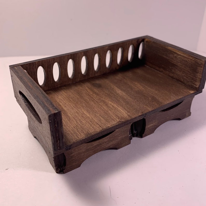 May include: A small, dark brown wooden tray with a rectangular shape. The tray has a series of oval cutouts along one side and curved cutouts on the ends. The wood grain is visible, giving it a natural look.