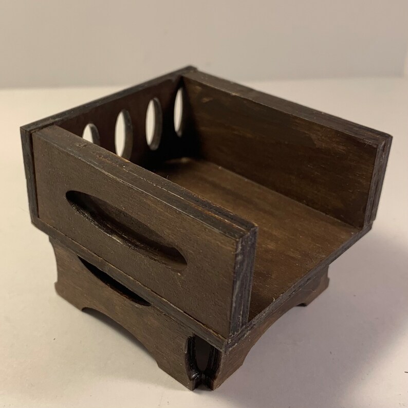 May include: A small, dark brown wooden box with a rectangular shape. The box has a slatted back with oval cutouts and a curved design on the front. The wood grain is visible, and the box appears to be handmade.