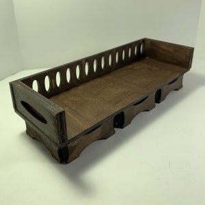 May include: A dark brown wooden tray with a rectangular shape. The tray has a series of oval cutouts along one side and decorative cutouts on the sides. The tray is designed to hold items.