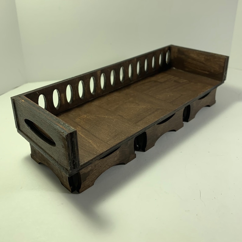 May include: A dark brown wooden tray with a rectangular shape. The tray has a series of oval cutouts along one side and decorative cutouts on the sides. The tray is designed to hold items.
