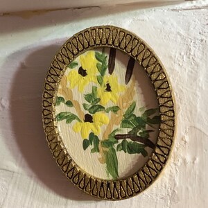 May include: A small, round, framed painting of yellow flowers with green leaves on a white background. The frame is gold with a black and gold patterned border.