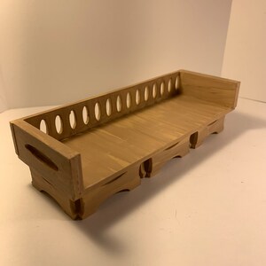 May include: A small, wooden decorative bench painted in a light brown color. The bench has a series of oval cutouts along the back and decorative cutouts on the sides. The bench is designed with a flat surface for display.