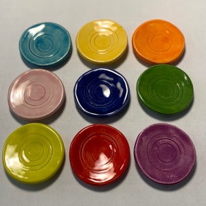 以下が含まれることがあります： 白い表面に、同心円模様の入った様々な色の小さなセラミック製の皿が9枚、3×3のグリッドパターンで並んでいます。