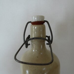 May include: A ceramic bottle with a metal wire closure and a white ceramic stopper. The bottle is beige and has a textured surface.