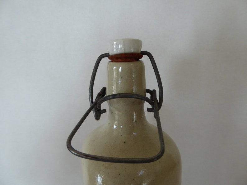 May include: A ceramic bottle with a metal wire closure and a white ceramic stopper. The bottle is beige and has a textured surface.