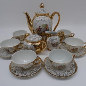 May include: A vintage porcelain tea set with gold trim. The set includes a teapot, sugar bowl, creamer, and several teacups with saucers. The white porcelain is decorated with a floral and pastoral scene.